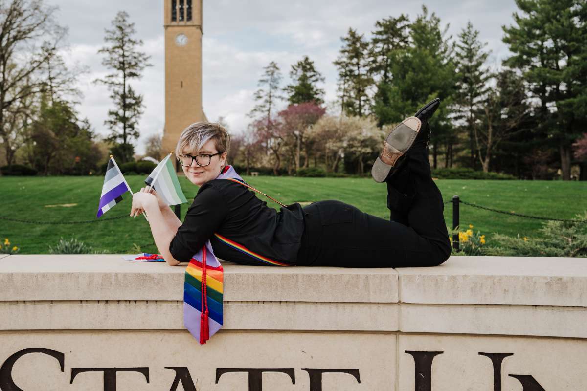 Student with Lavender Graduation Celebration Stole, Asexual Flag, and Aromantic Flag. Student is laying on the Iowa State Sign behind the Memorial Union.