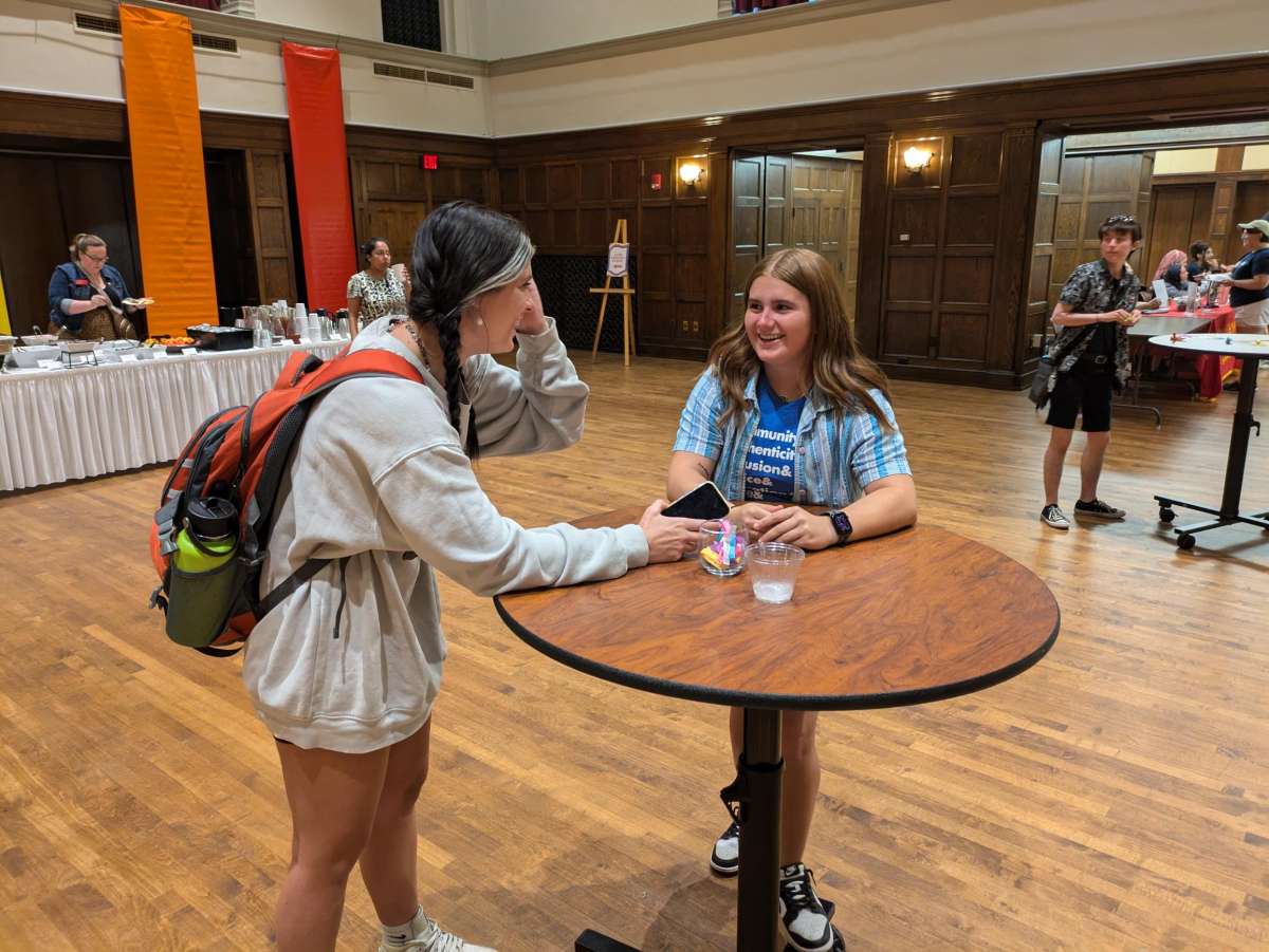 Two students talking at a cocktail table at Community Welcome. One student wearing a backpack. Both students smiling and laughing