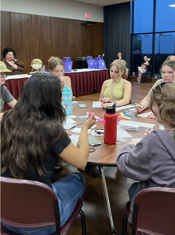 Students sitting around a table with bingo cards. Drag queen Coco announcing drag numbers with golden bingo cage in background.