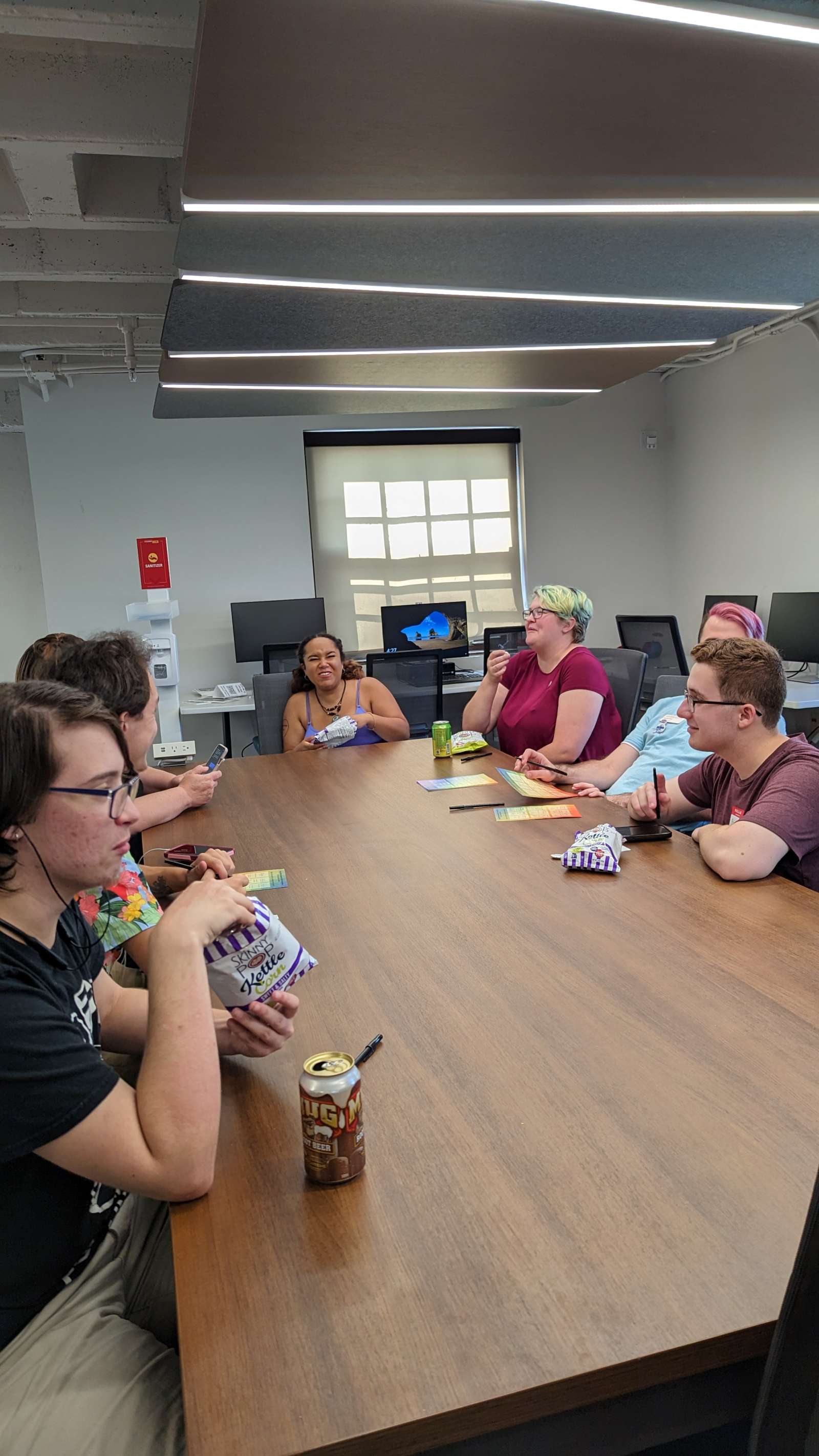 Students sitting around the large table at the Center for LGBTQIA+ Student Success. Many students conversing and laughing while eating snacks and playing Human Bingo.
