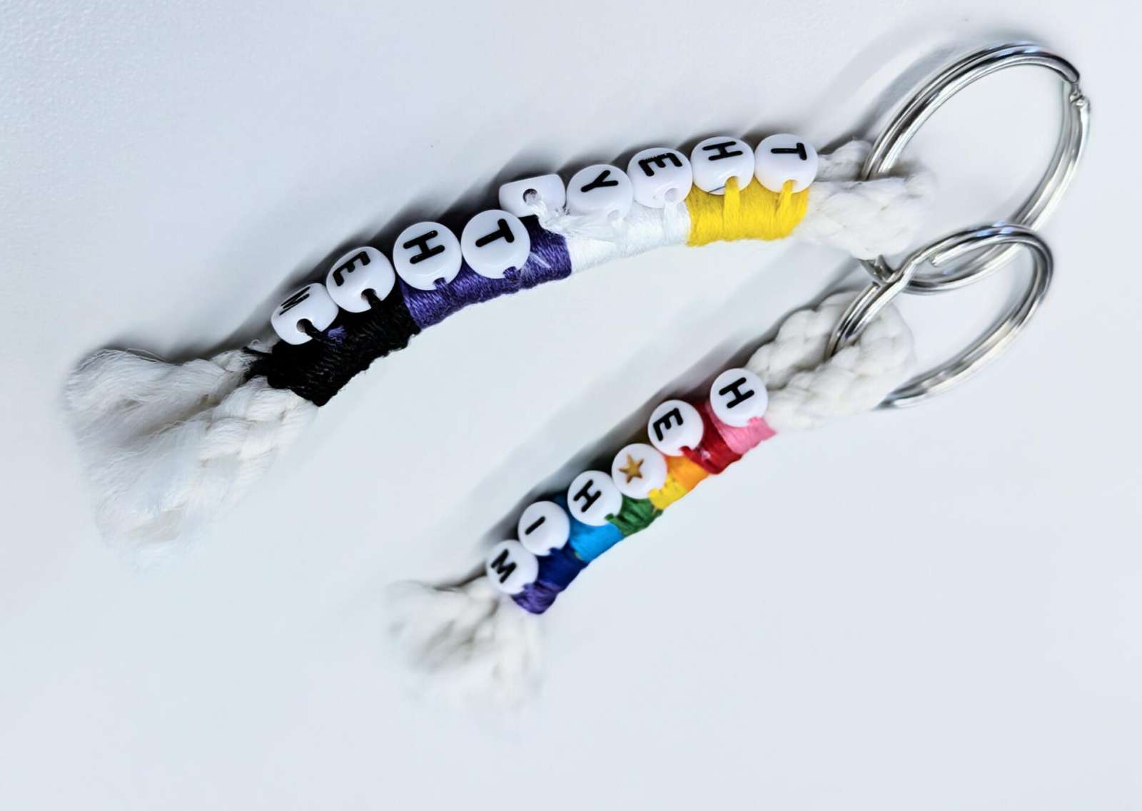 Two keychains. One keychain in the colors of the nonbinary pride flag: yellow, white, purple, and black; with beads spelling "they/them" Another keychain in the colors of the Gilbert Baker Pride Flag: pink, red, orange, yellow, green, turquoise, indigo, violet; with beads spelling "he/him"